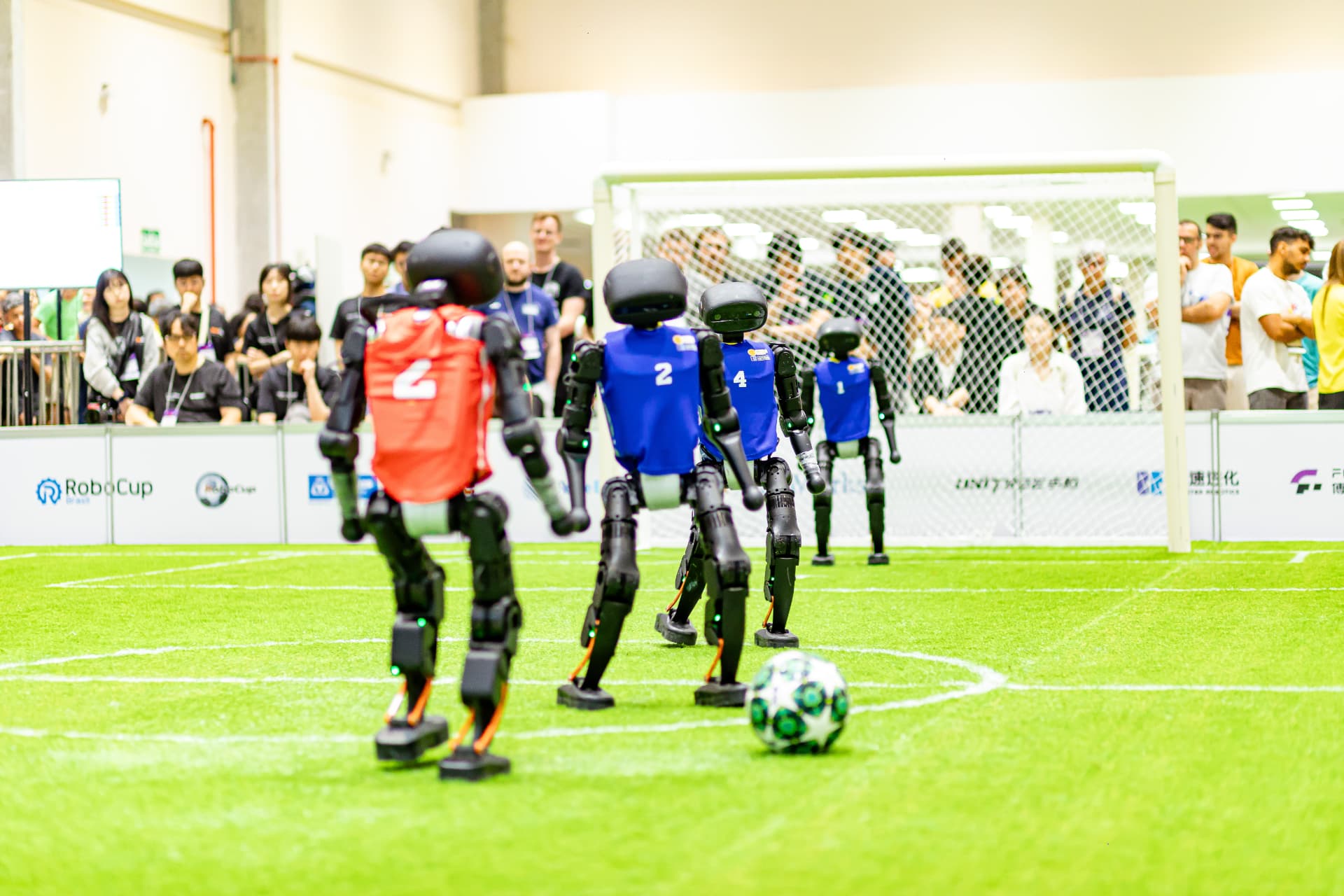 Robôs em fila olhando a bola, com a torcida ao fundo