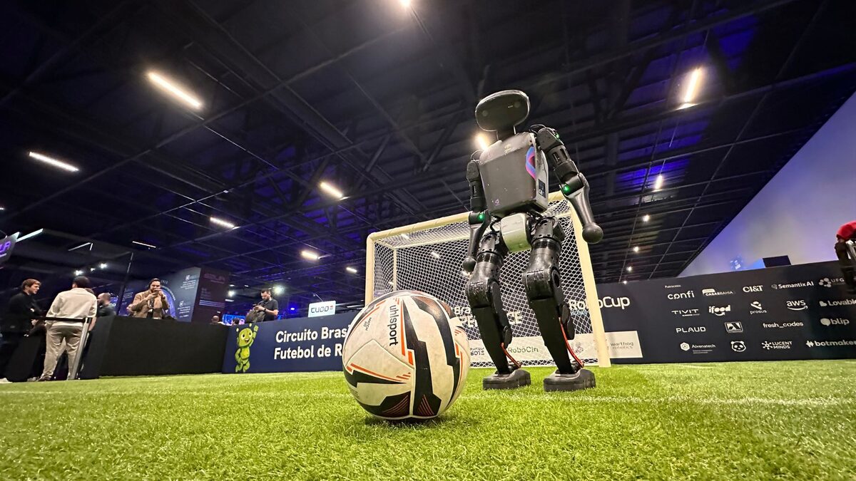 Robô ao lado da bola e do gol, visto de baixo, com o telão e os patrocinadores ao fundo